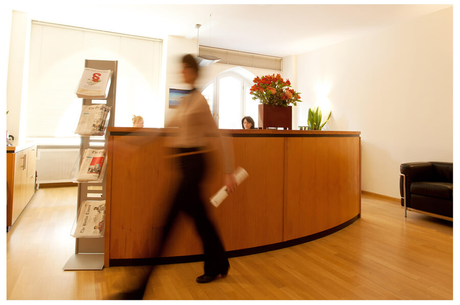 Eine Frau mit einem eleganten Kleid geht vor einem Empfangstresen, an dem ein Empfangsmitarbeiter steht, entlang