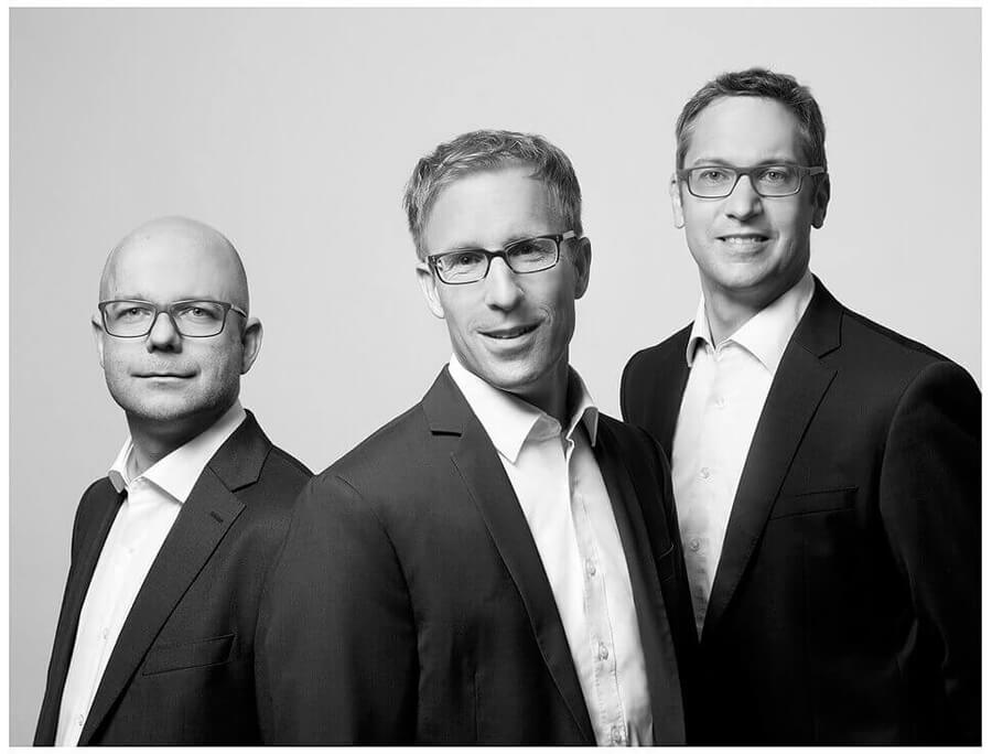 Drei Geschäftsführer in Anzügen und Brillen ohne Krawatten haben sich für ein schwarz-weiß Foto arrangiert und wirken wie ein Team