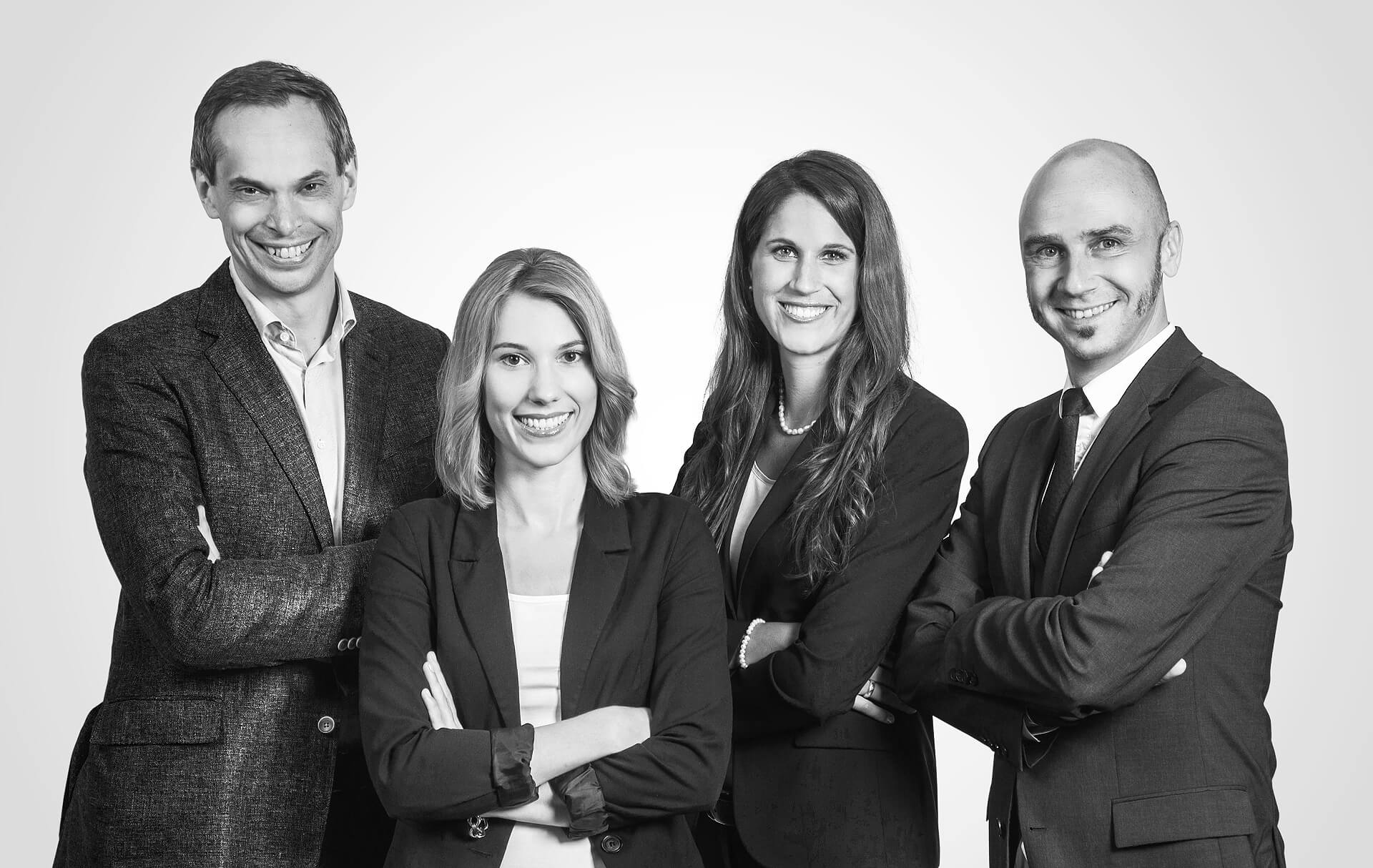 Ein schwarz-weißes Foto zeigt eine Gruppe von Geschäftsleuten, die in einer Reihe stehen. Sie lächeln in die Kamera und tragen elegante Kleidung. Der Hintergrund ist unscharf