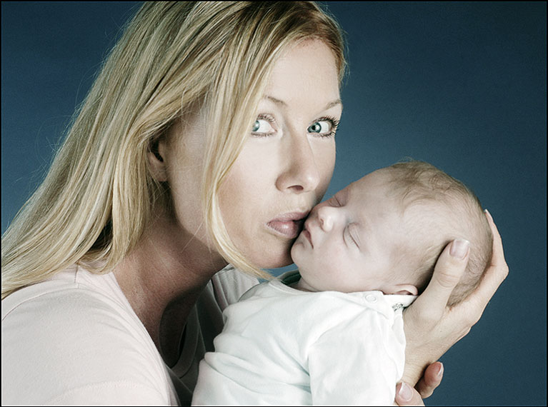 Portrait einer Mama mit dem Neugeborenen Baby in der Hand, schauend in die Kamera und küsst das Baby