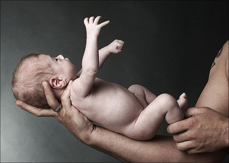 Eine tätowierte Männerhand hält ein Baby in die Luft mit den beiden Händen