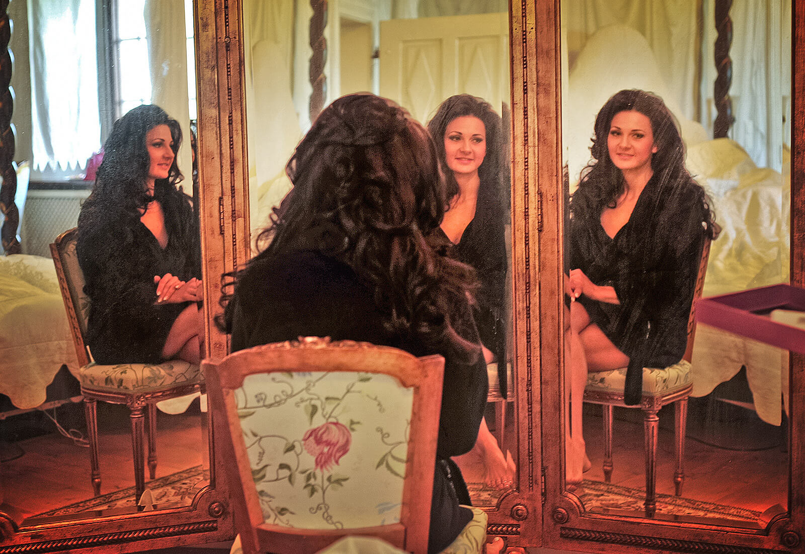 Braut mit langen, dunklen Haaren sitzt vor einem antiken Dreier Spiegel. Sie betrachtet ihr Spiegelbild und lächelt zufrieden