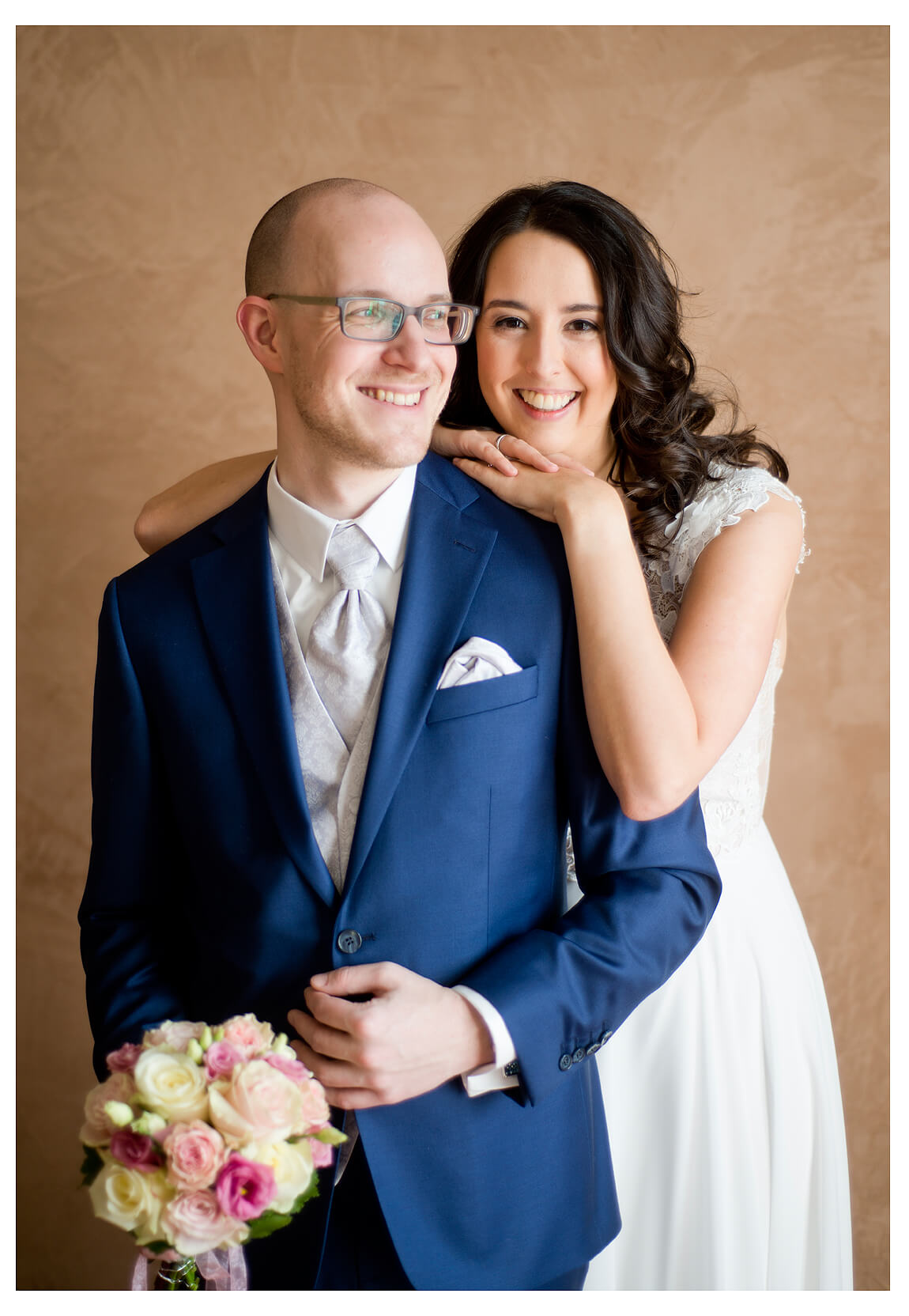 Ein glückliches Brautpaar posiert für ein Foto vor einer braunen Wand, Bräutigam hält Blumenstrauss in der rechten Hand