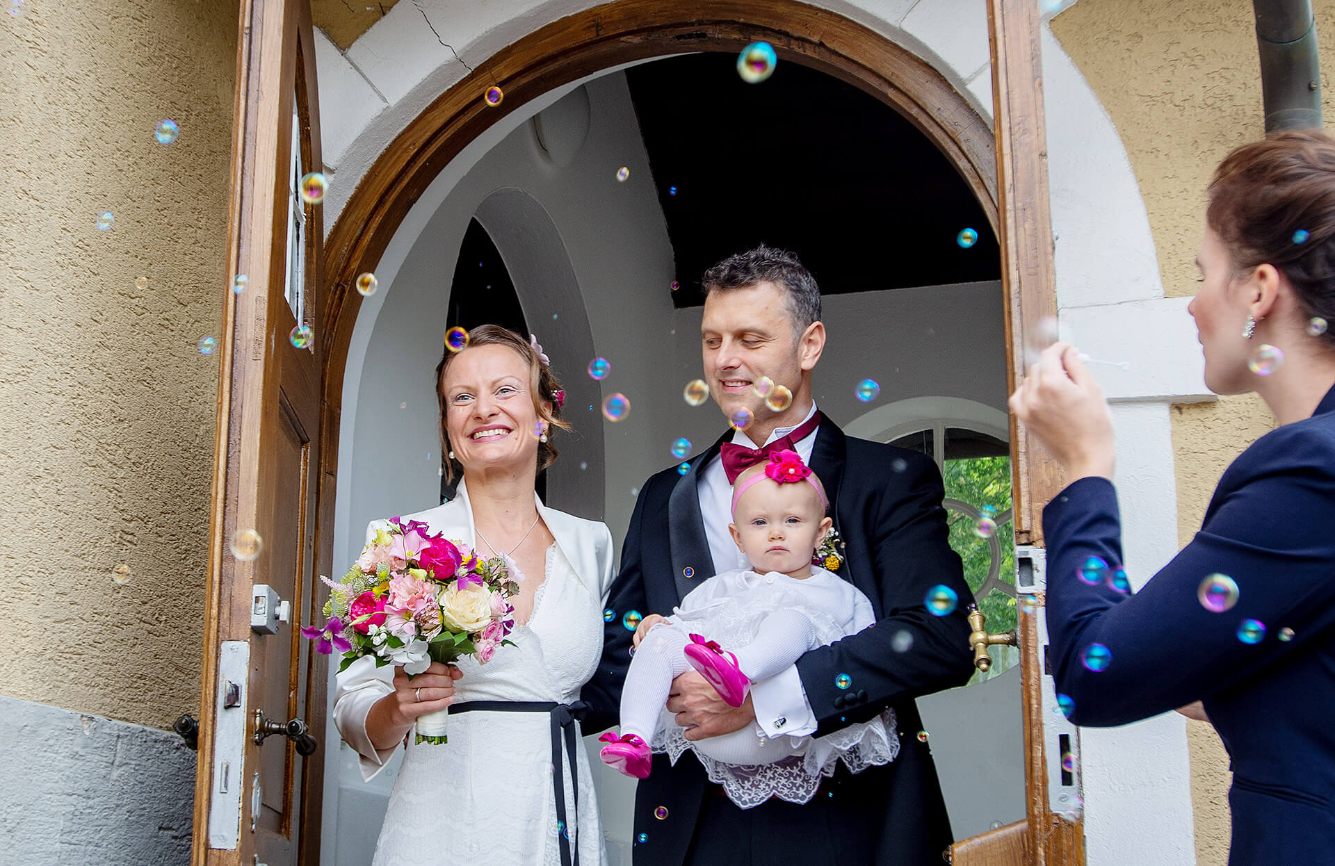 Eine wunderschöne Braut und ein eleganter Bräutigam und einem Baby auf dem Arm stehen vor einer mit Blumen geschmückten Tür und umgeben von schwebenden Seifenblasen