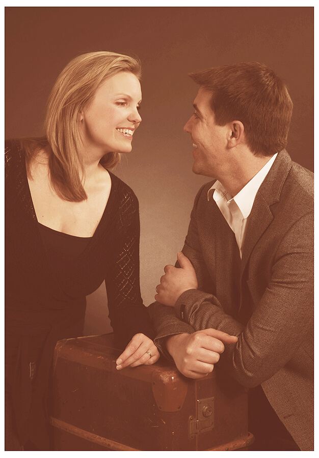 Ein Paar, bestehend aus einem Mann und einer Frau, sitzt auf einem Koffer und lässt sich für ein Fotoshooting ablichten, schaut lächelnd zueinander