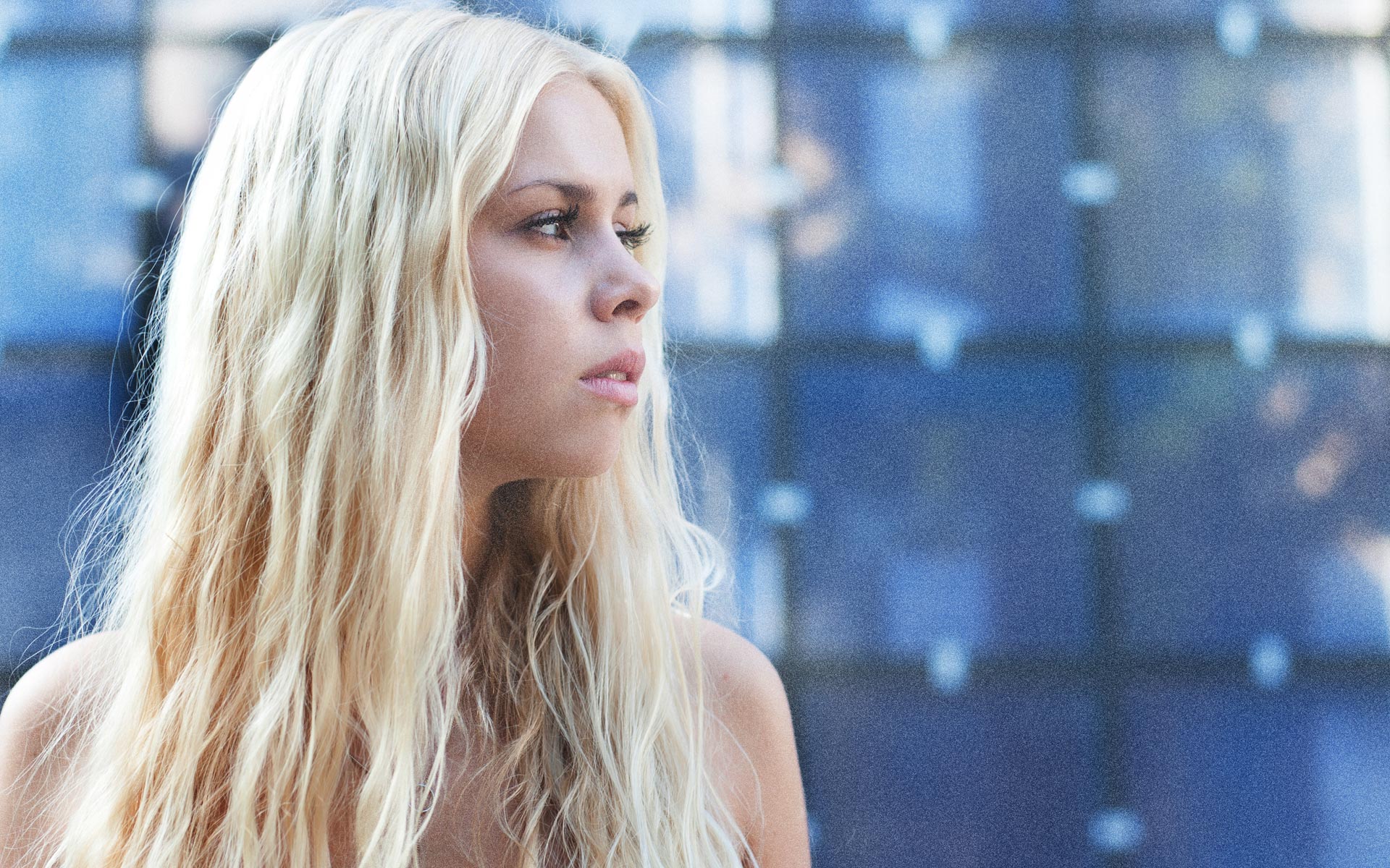 Eine attraktive blonde Frau mit langen Haaren posiert vor einer blauen Wand. Sie schaut zur Seite, eine selbstbewusste Ausstrahlung.