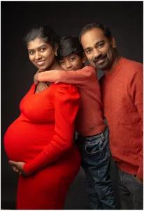 Eine schwangere Frau in einem leuchtend roten, eng anliegenden Kleid steht links vor einem dunklen Hintergrund. Ihr Babybauch ist deutlich zu sehen, während sie lächelnd in die Kamera schaut. Rechts neben ihr steht ein Mann in einem roten Pullover, der ebenfalls lächelt. Zwischen den beiden, leicht erhöht, legt ein Junge in einem roten Oberteil seine Arme liebevoll auf die Schultern seiner Mutter und schaut dabei über ihre Schulter in die Kamera. Alle drei sind farblich aufeinander abgestimmt und strahlen eine warme, familiäre Verbundenheit aus
