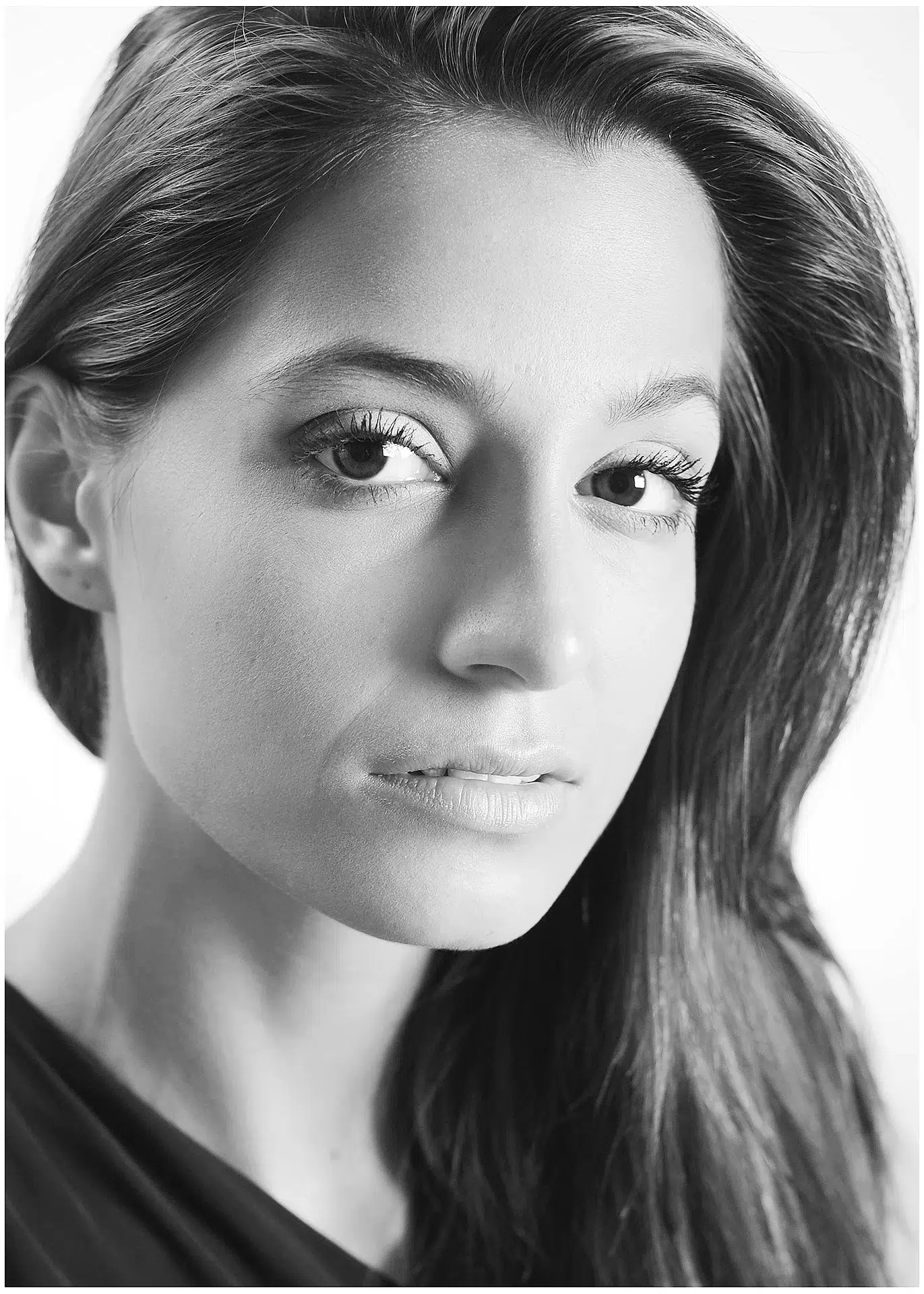 Jenefer Riili in einer klassischen Schwarz-Weiß-Nahaufnahme. Ihr Gesicht ist leicht zur Seite geneigt, während sie mit ausdrucksstarken Augen und weichen Gesichtszügen in die Kamera schaut. Der Hintergrund ist hell, wodurch ihr Gesicht besonders betont wird.