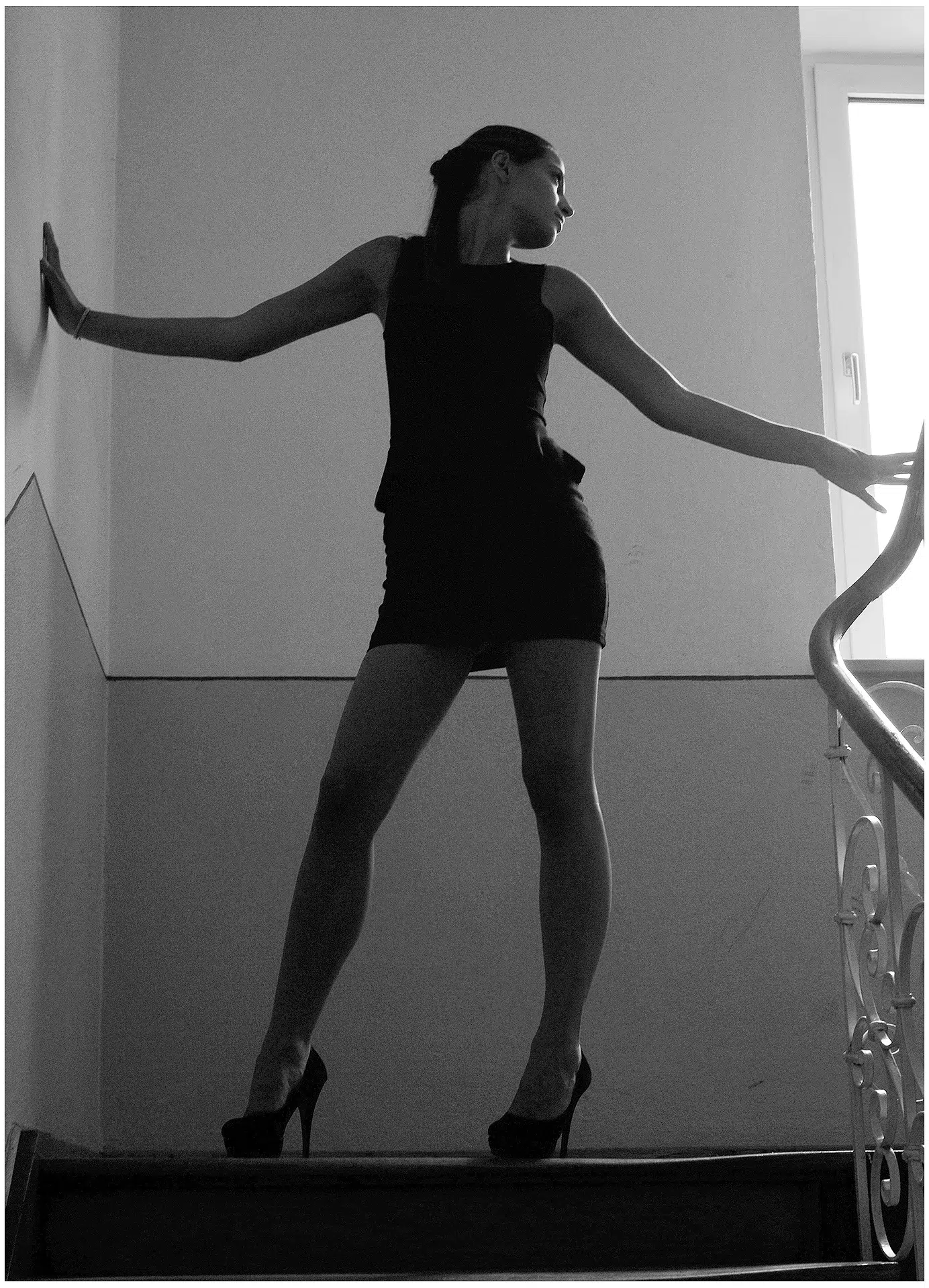 Jenefer Riili in einer dramatischen Schwarz-Weiß-Aufnahme. Sie steht auf einer Treppe, ihre Silhouette hebt sich gegen das Licht aus einem Fenster ab. Ihre Pose mit ausgestreckten Armen erzeugt eine elegante, fast skulpturale Wirkung.

