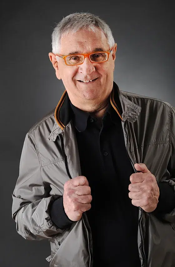 Ein älterer Herr mit silbergrauem Haar und markanten Gesichtszügen trägt eine sportlich geschnittene, graue Jacke mit dunklem Innenfutter und ein schwarzes Polo-Shirt. Seine orangefarbene Brille verleiht ihm eine moderne, stilbewusste Note. Er hält die Jacke mit beiden Händen leicht geöffnet und lächelt herzlich. Der dunkle Studiohintergrund unterstreicht die Dynamik seines lässigen, selbstbewussten Auftretens.

