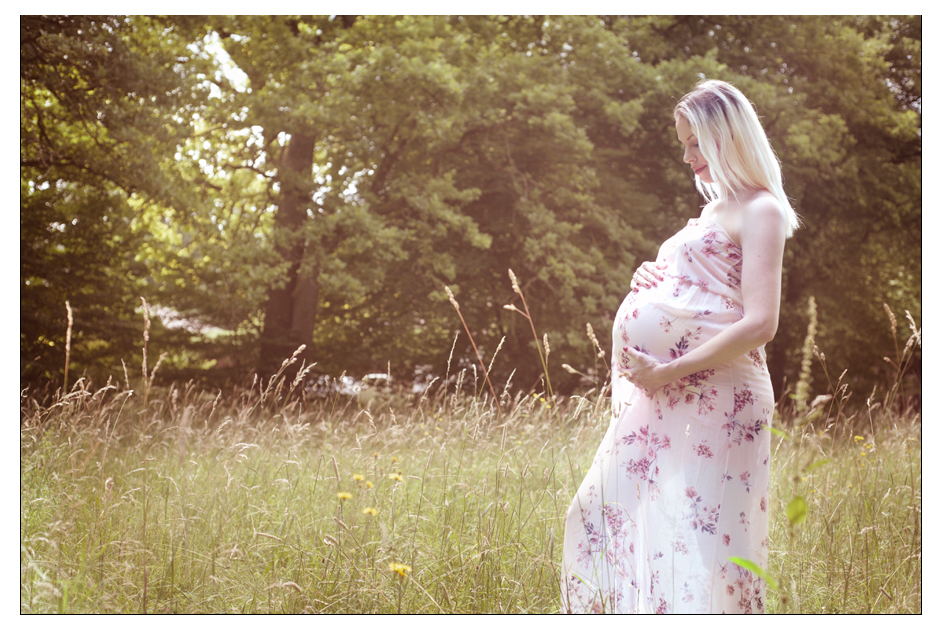 Babybauch-Shooting in Grünwald bei München mit einer schwangeren Frau im rosa Blumenkleid auf einer sonnigen Sommerwiese