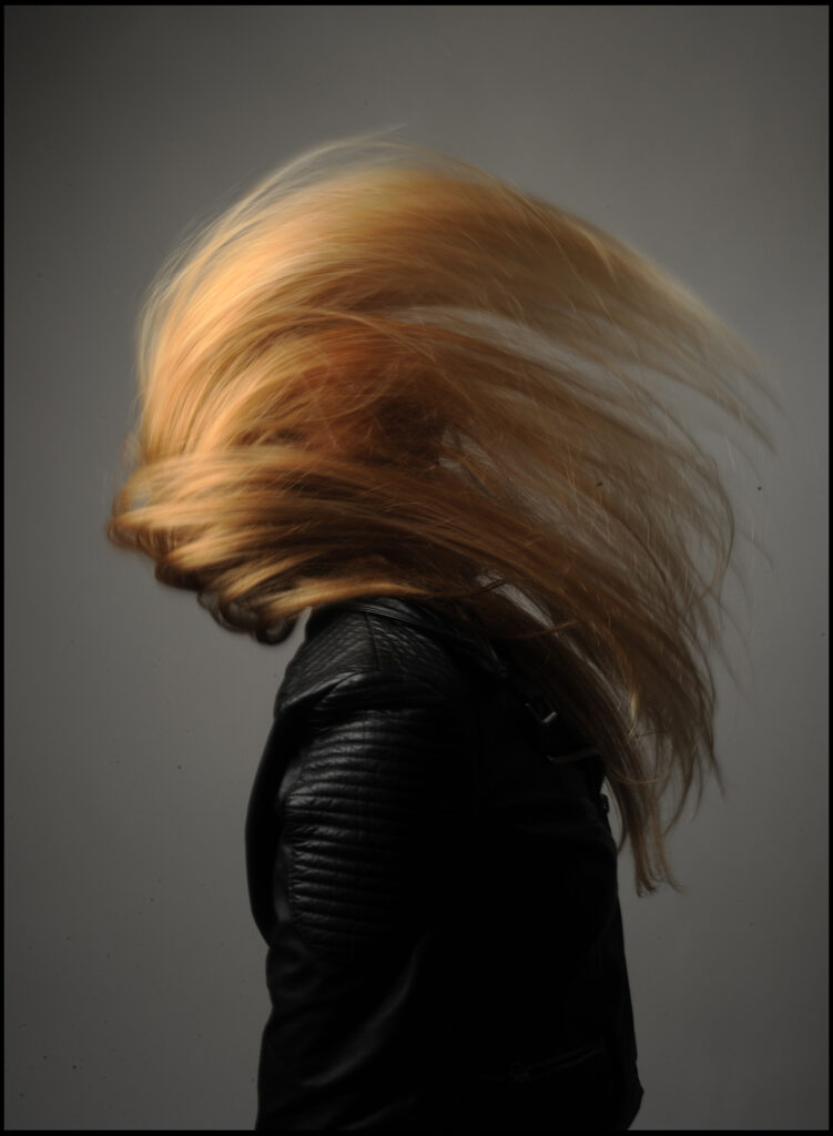 Beauty Fotoshooting im Fotostudio Belichtungswert München, junge blonde Frau mit langem Haar in Bewegung, schwarze Lederjacke und dynamische Fashion-Pose im Studio
