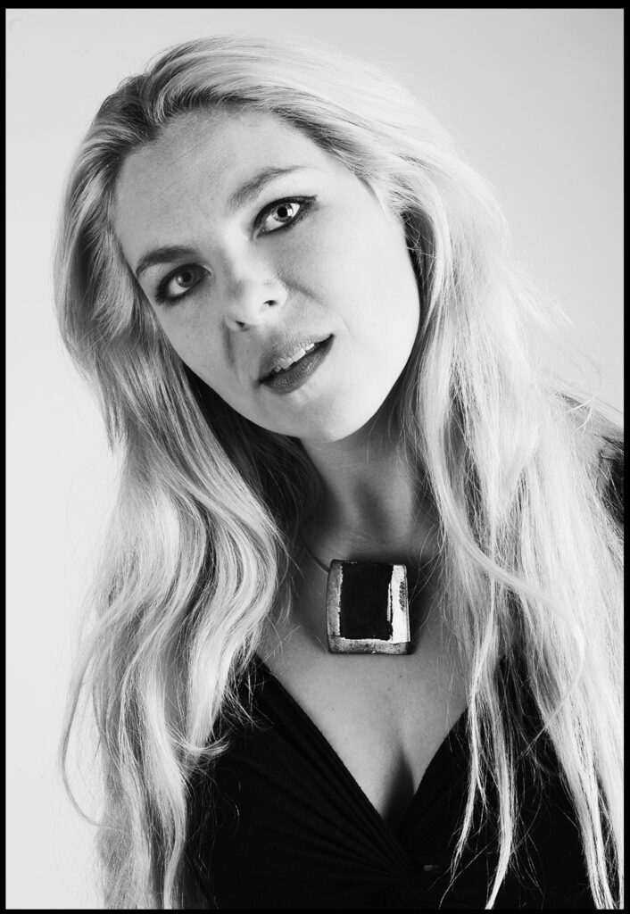Beauty Fotoshooting im Fotostudio Belichtungswert München, junge blonde Frau als Schwarzweiß-Portrait mit langen Haaren, dunklem Outfit, markanter Kette und dramatischem Blick