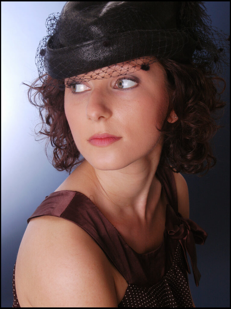 Beauty-Fotoshooting im Fotostudio Belichtungswert München, junge Frau mit schwarzem Hut, weichem seitlichem Blick, dunklem Kleid und ruhiger Portrait-Komposition