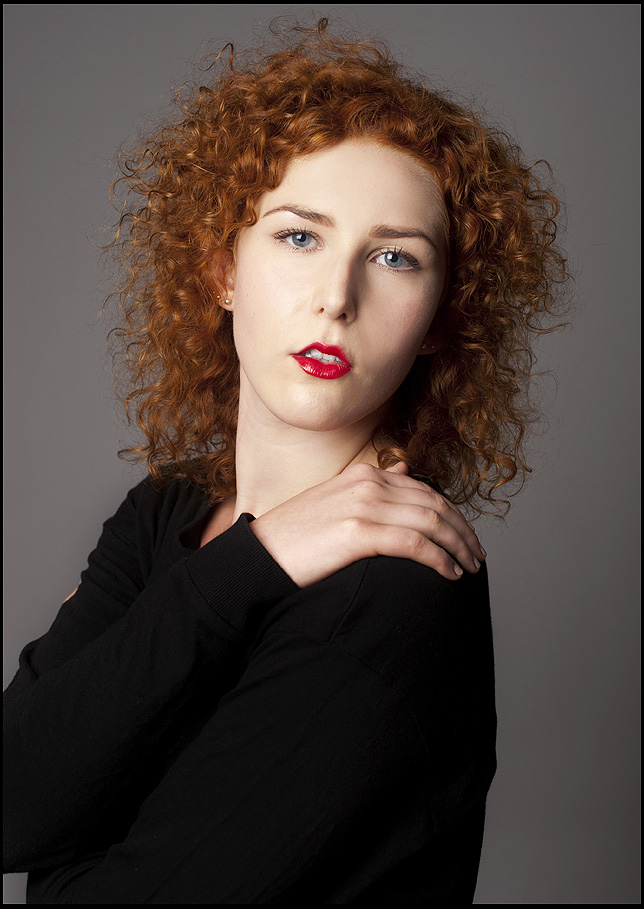 Beauty-Fotoshooting im Fotostudio Belichtungswert München, junge rothaarige Frau mit lockigem Haar, schwarzem Oberteil, rotem Lippenstift und direktem dramatischem Blick