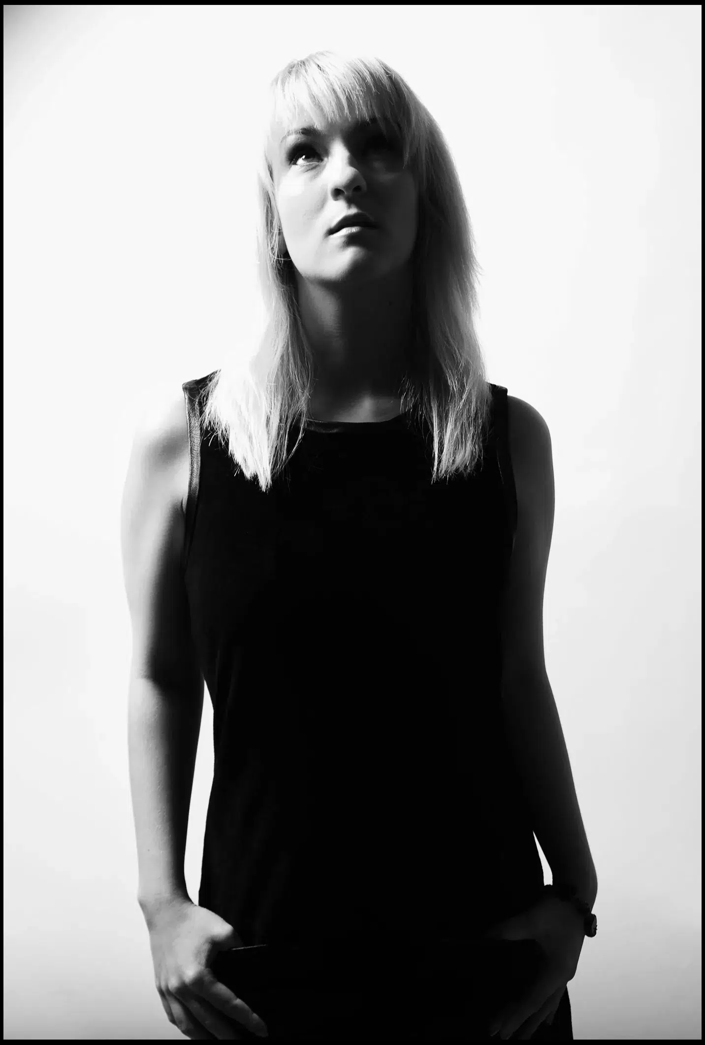 Schwarzweißfoto einer Person mit hellem Haar, die ein ärmelloses, dunkles Oberteil trägt und vor einem schlichten weißen Hintergrund leicht nach oben blickt. Die Schönheit des Fotoshootings wird in ihrem heiteren Ausdruck eingefangen.
