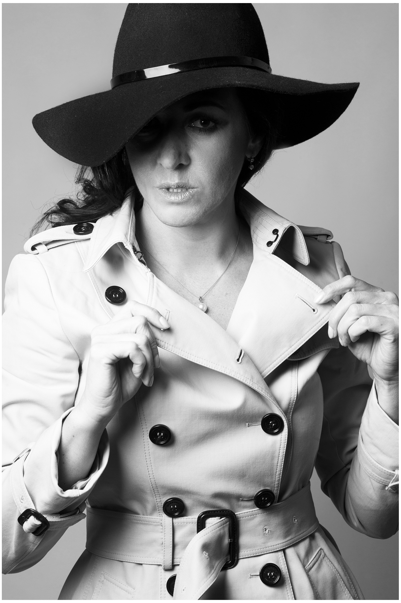Best-Age Model Sedcard vom Fotostudio Belichtungswert München, Schwarzweiß-Fashion-Portrait einer Frau mit schwarzem Hut und hellem Trenchcoat, die den Kragen mit beiden Händen hält und direkt in die Kamera blickt