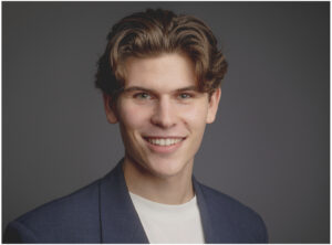 Professionelles Bewerbungsfoto eines jungen Mannes im Business-Casual-Look mit dunkelblauem Blazer und weißem Shirt vor grauem Studiohintergrund in München.