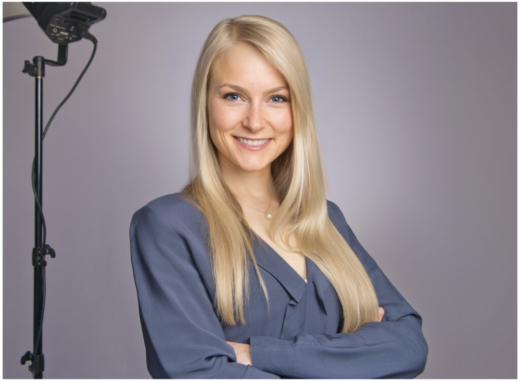 Bewerbungsfoto vom Fotostudio Belichtungswert München, blonde Frau in dunkelblauer Bluse lächelt freundlich mit verschränkten Armen vor grauem Studiohintergrund mit sichtbarer Softbox links.
