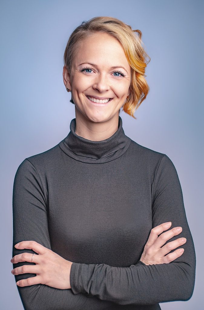 Modernes Bewerbungsfoto einer sympathisch lächelnden Frau im dunklen Rollkragen, fotografiert mit verschränkten Armen vor hellblauem Hintergrund