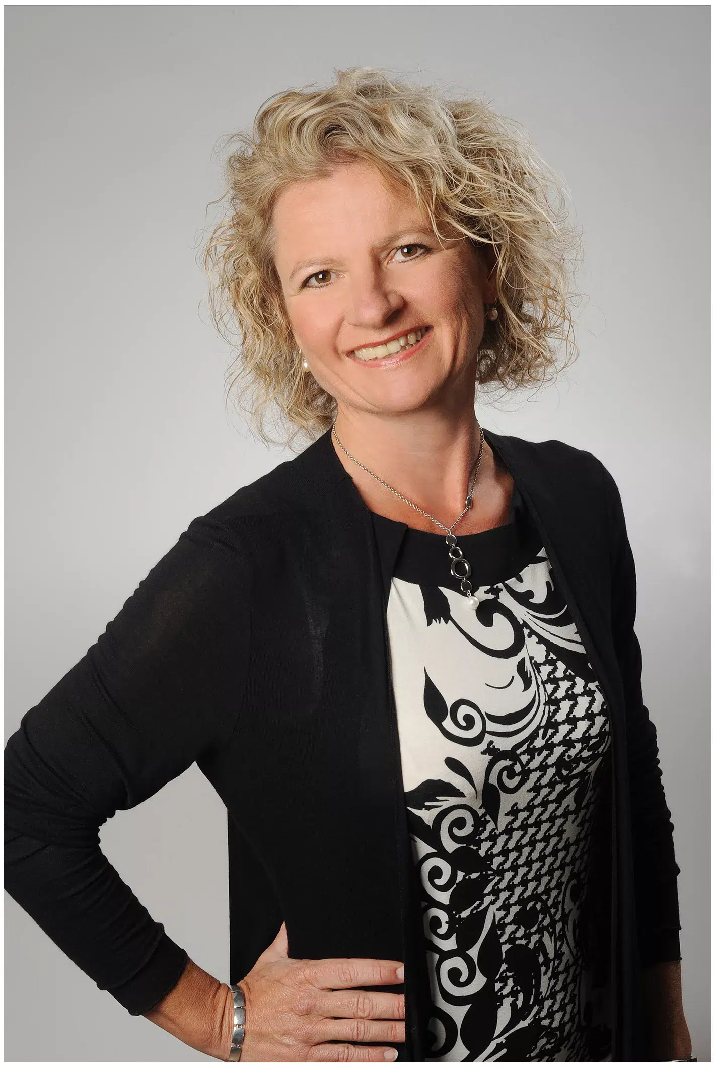 Eine Frau mit lockigem blondem Haar lächelt in die Kamera. Sie trägt eine schwarze Strickjacke über einem gemusterten Kleid und hat ihre Hand auf der Hüfte. Der schlichte graue Hintergrund unterstreicht den professionellen Look, perfekt für Bewerbungsfotos München