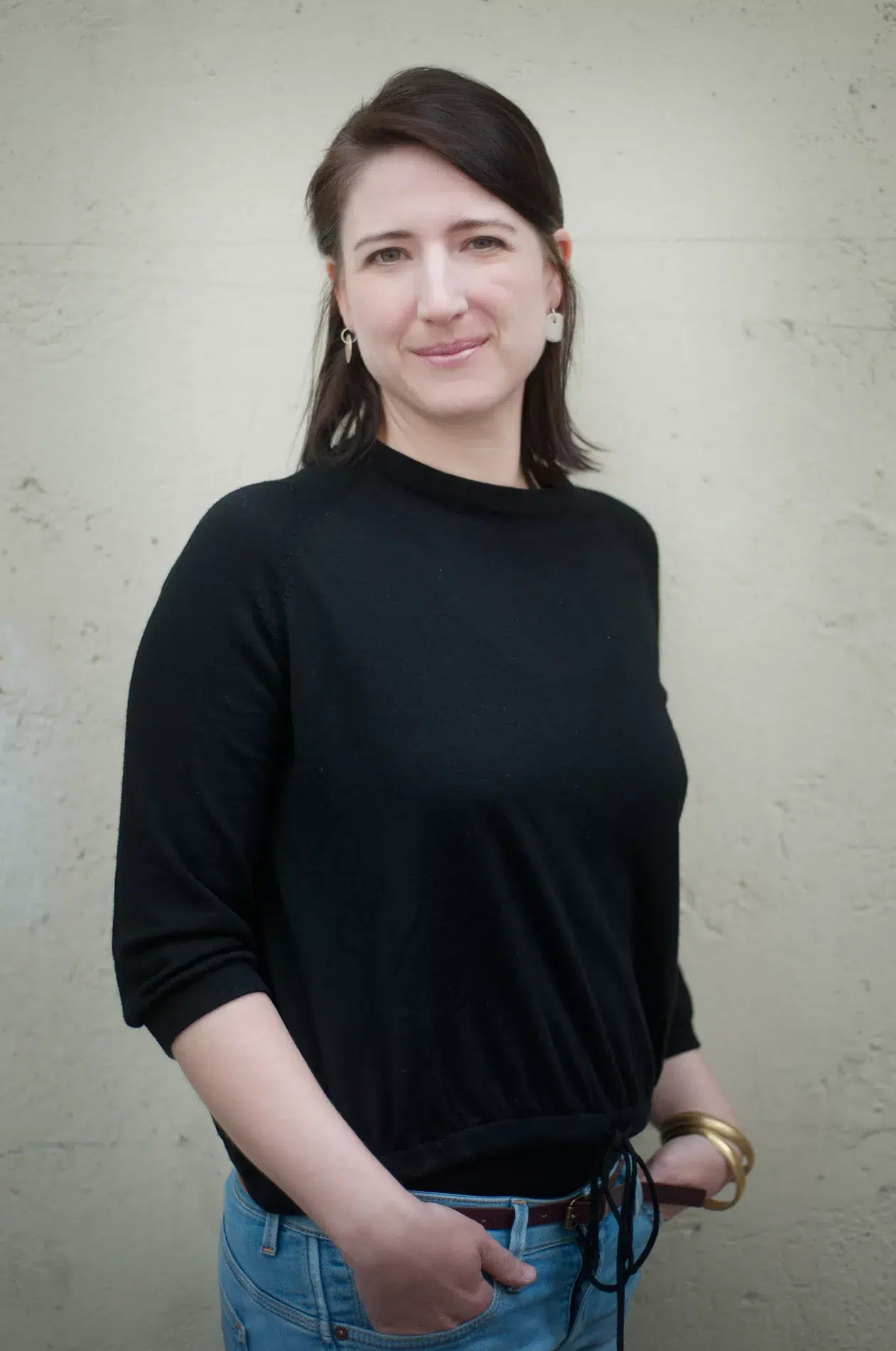 Eine Frau in schwarzer Bluse und Jeans steht vor einer Wand. Ihre Kleidung und Pose sind besonders betont.

