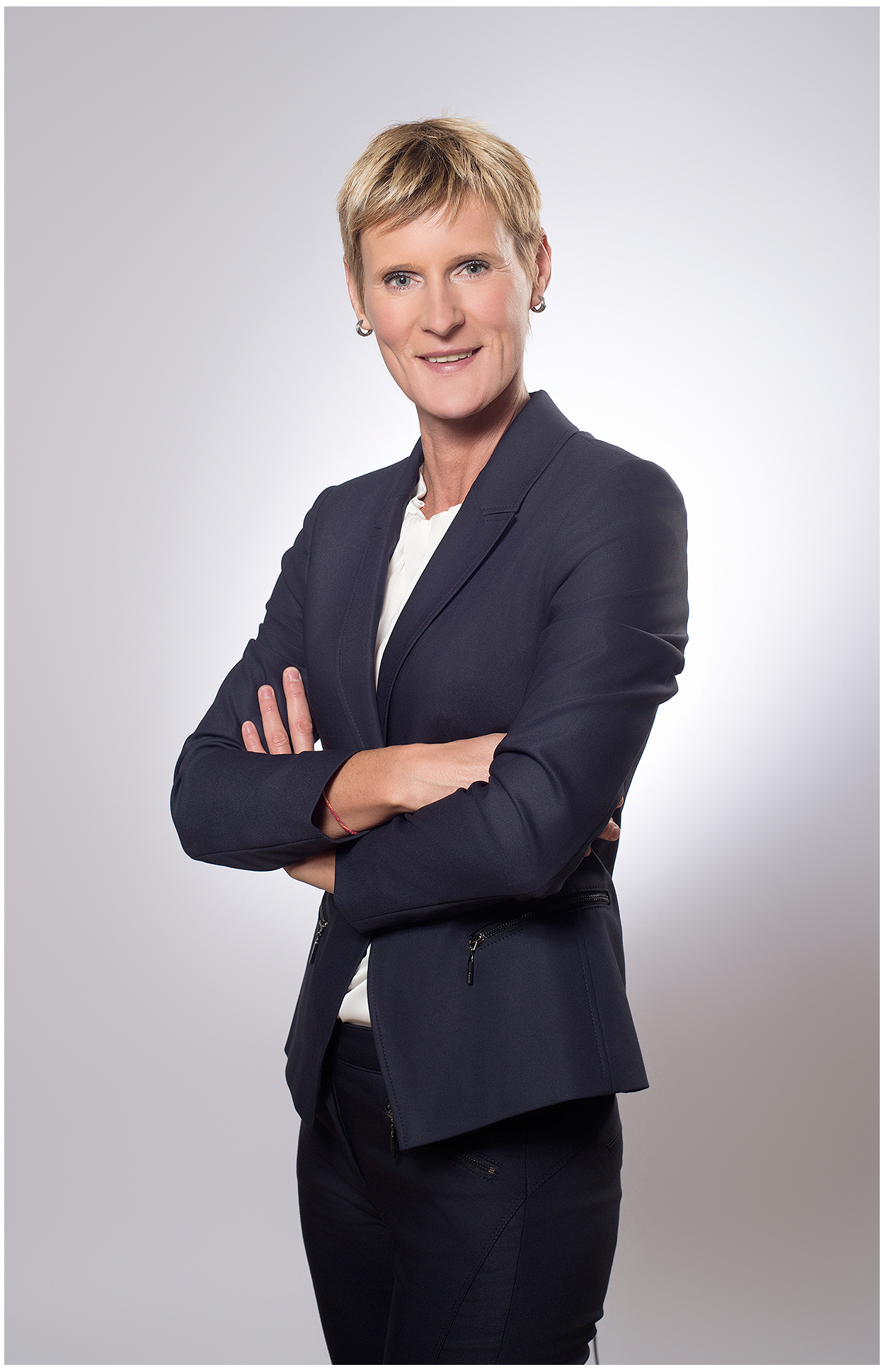 beschreibung: businessfoto-im-fotostudio-frau-im-dunkelblauen-hosenanzug-mit-weisser-bluse-arme-verschränkt-selbstbewusste-pose-vor-hellem-hintergrund