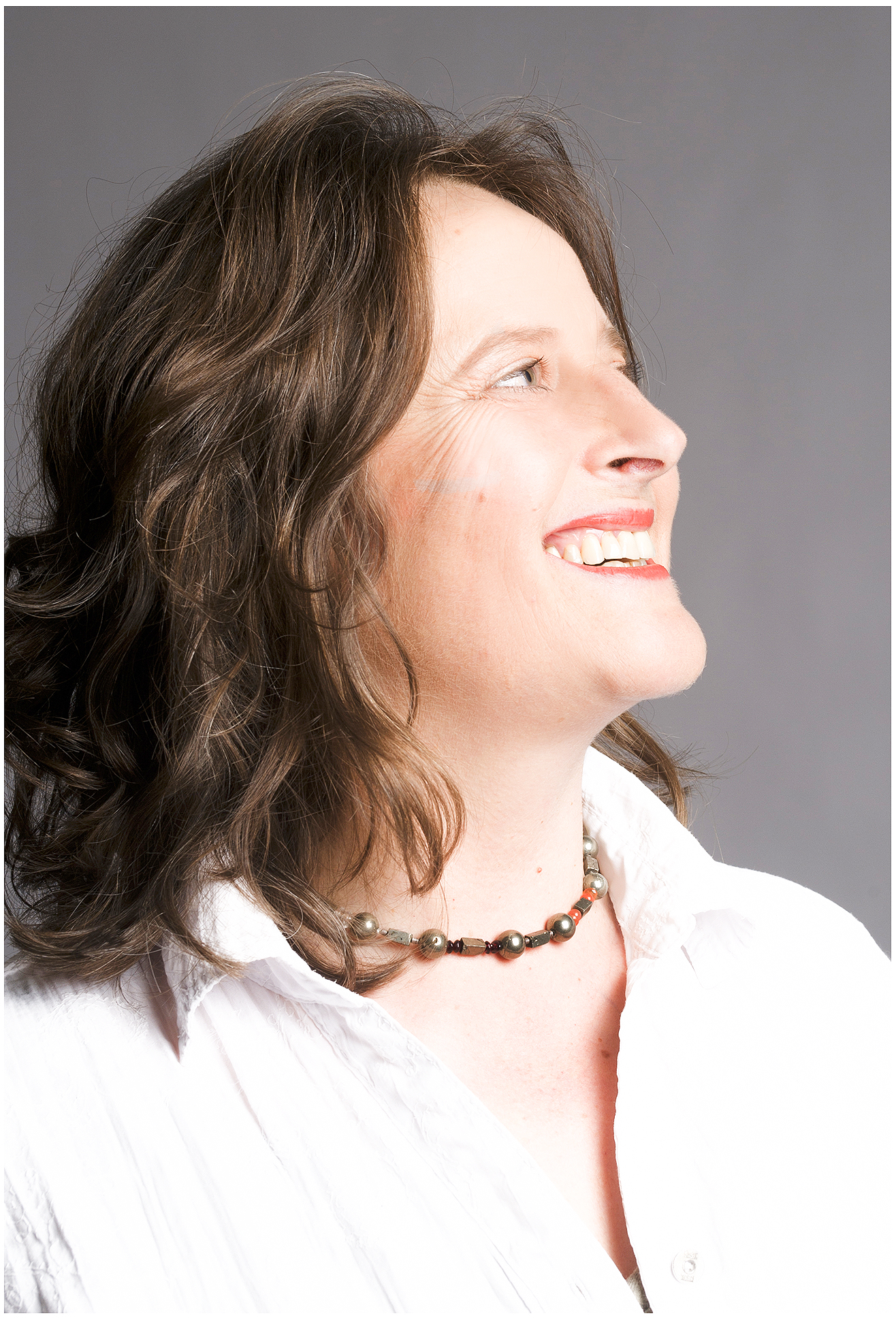 Nahes Profil-Businessportrait einer Coachin: seitlicher Blick, warmes Lachen, weiße Bluse vor grauem Hintergrund – Personal Branding Shooting München