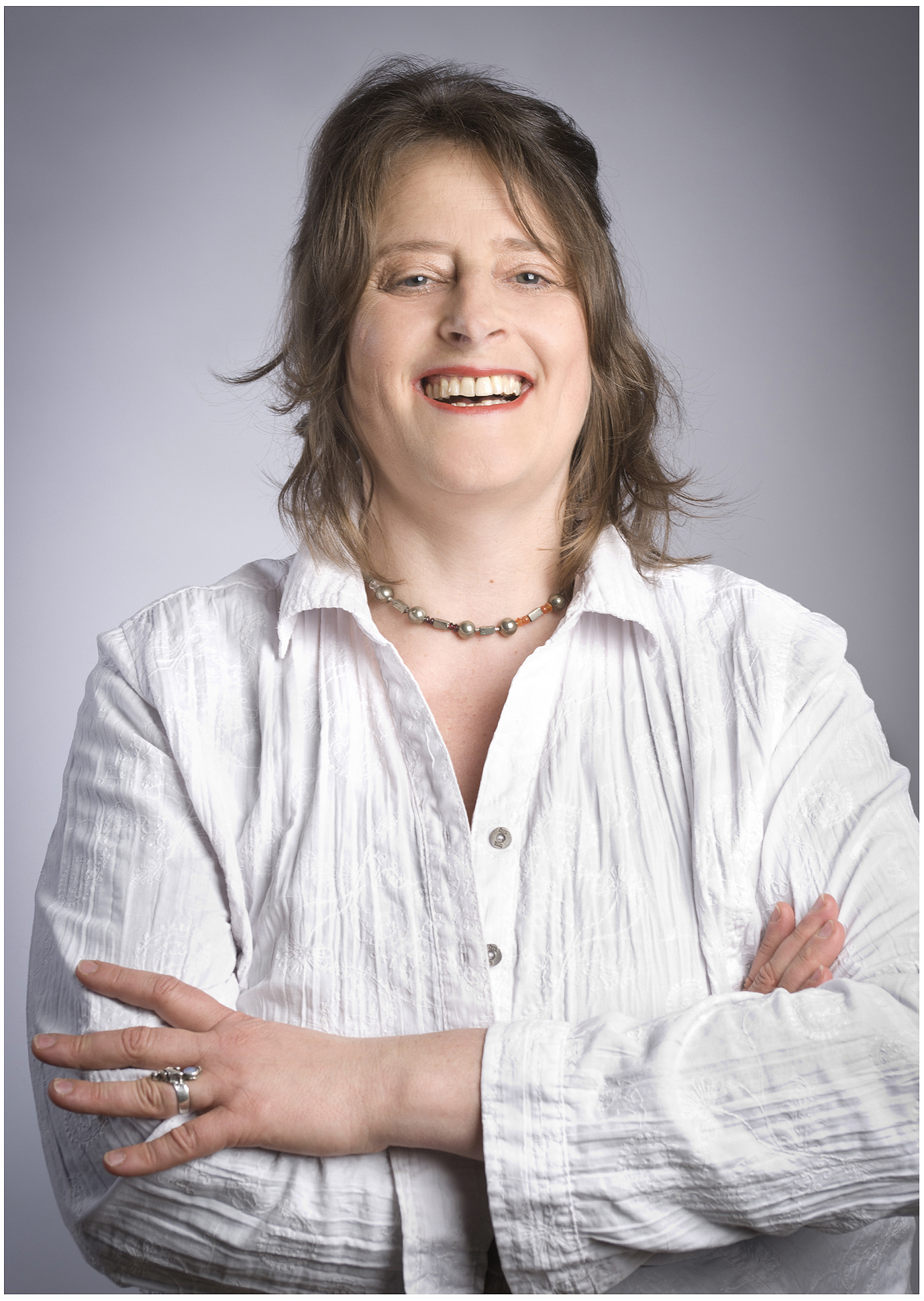 Businessportrait einer Coachin: weiße Bluse, Arme verschränkt, offenes Lachen vor grauem Hintergrund – authentische Firmenfotografie München