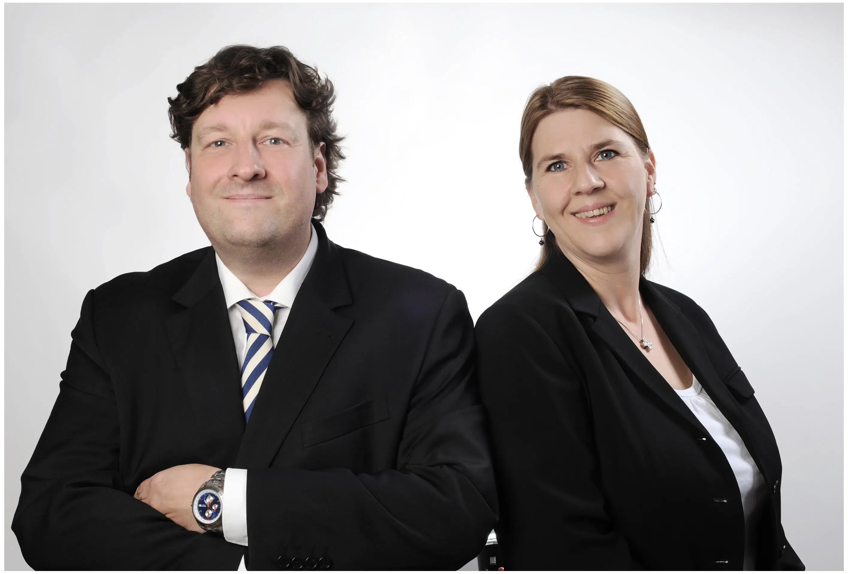 Ein professionelles Business-Portrait von zwei Dolmetschern: Ein Mann in einem schwarzen Anzug mit einer blau-weiß gestreiften Krawatte und eine Frau in einer schwarzen Blazer-Jacke mit einem weißen Oberteil darunter. Beide sitzen nebeneinander und blicken freundlich in die Kamera. Der Mann hat dunkle, leicht wellige Haare und eine silberne Armbanduhr am linken Handgelenk. Die Frau trägt eine silberne Kette und Ohrringe. Der Hintergrund ist neutral weiß