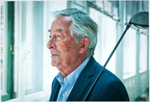 Portrait eines älteren Mannes im blauen Sakko im Profil, der einen Golfschläger über der Schulter trägt, fotografiert in heller Business-Umgebung