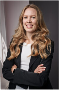 Portrait einer lächelnden jungen Businessfrau mit langen blonden Haaren, im schwarzen Blazer und weißem Top, mit verschränkten Armen in professioneller Studioatmosphäre