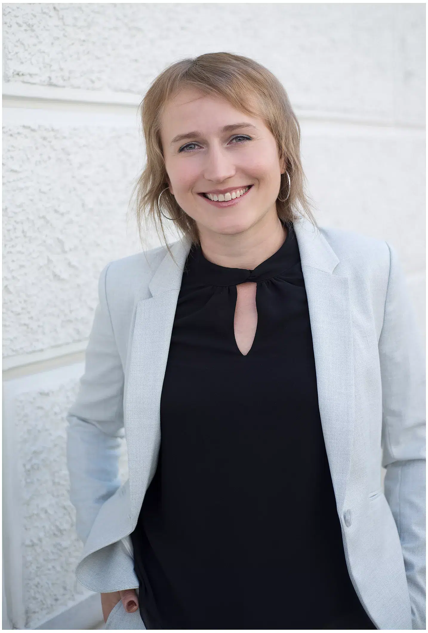 Junge Frau im hellen Blazer und schwarzem Oberteil draussen beim Fotoshooting graue Wand