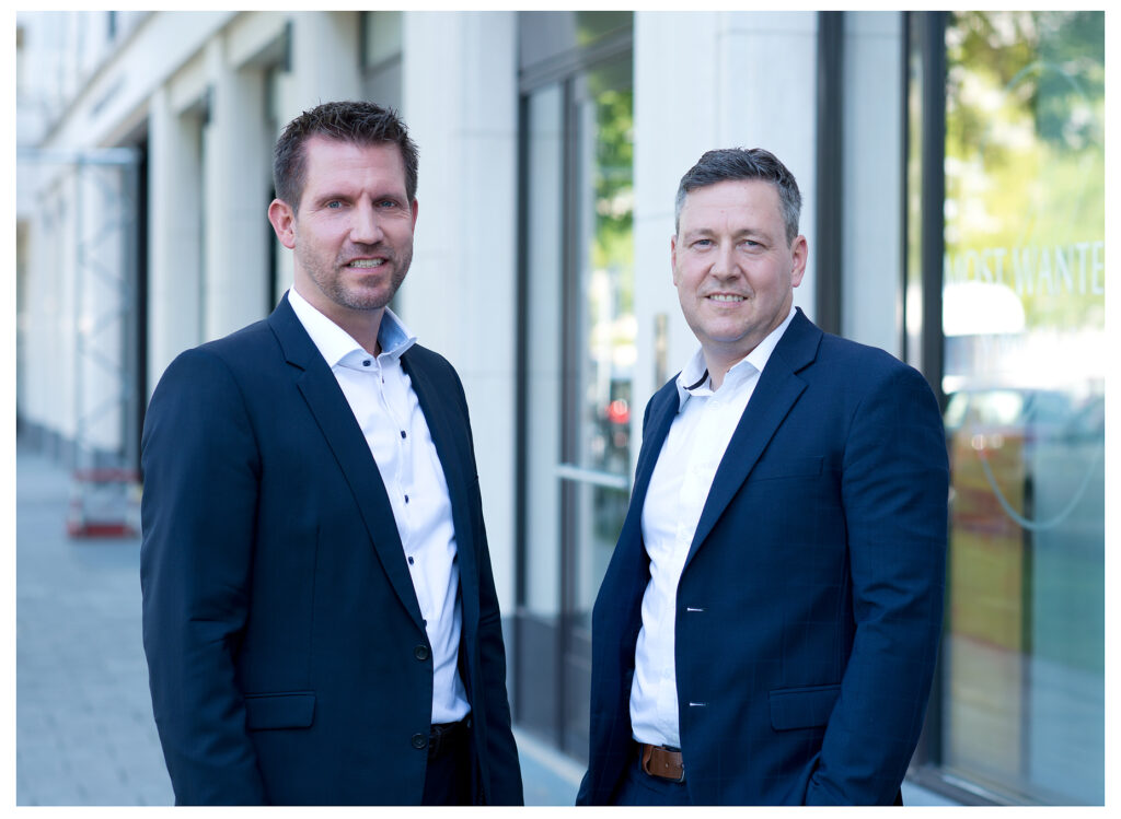Firmenshooting vom Fotostudio Belichtungswert München, zwei Männer im Anzug stehen vor moderner Firmenfassade für professionelle Outdoor Businessportraits