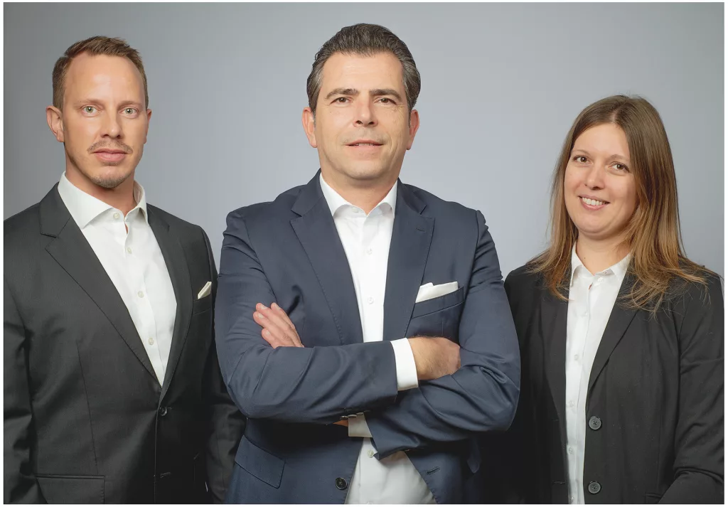 Firmenshooting mit professionellem Gruppenportrait von zwei Männern und einer Frau im Business-Outfit vor hellgrauem Hintergrund im Fotostudio München