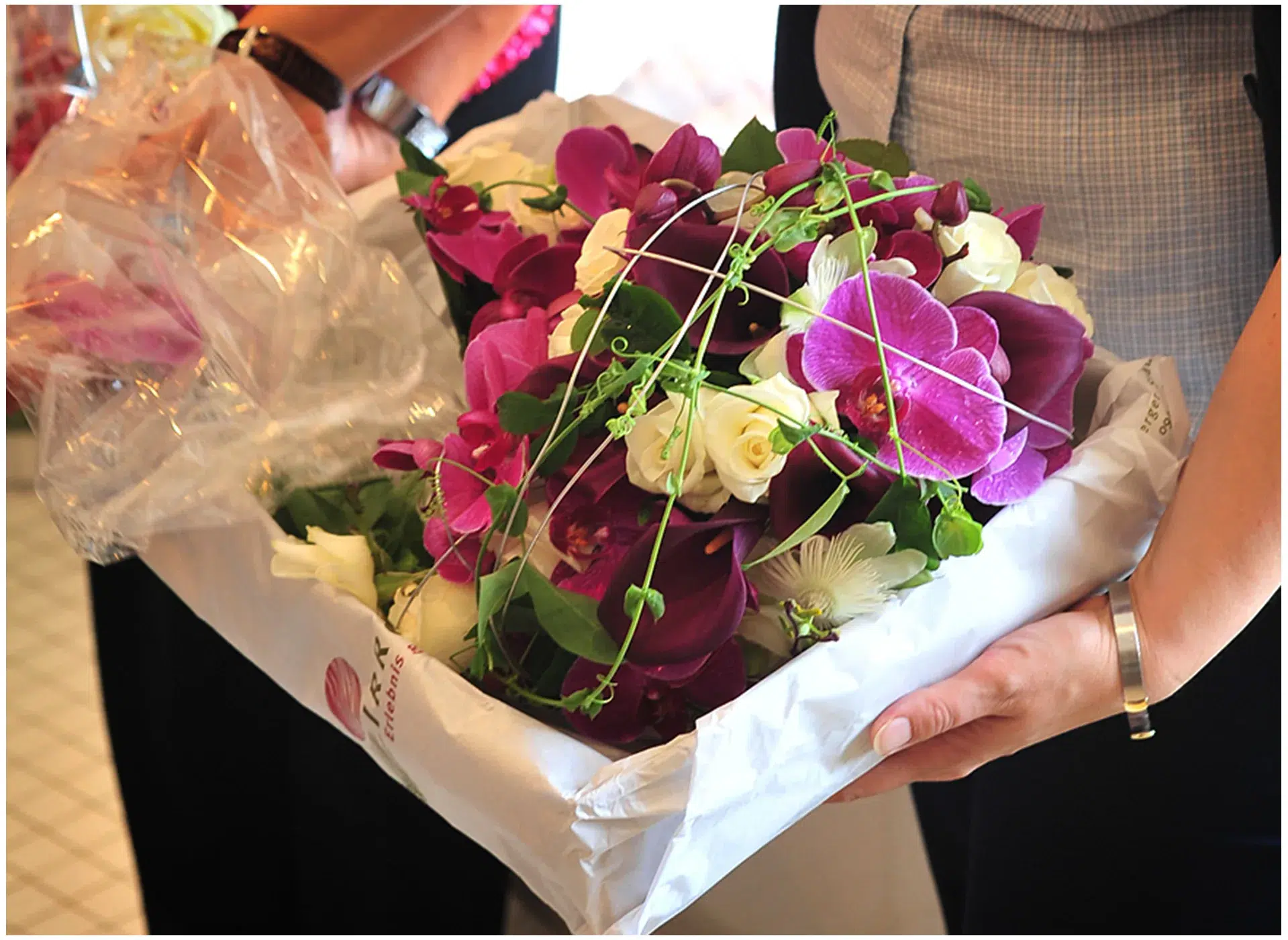 Eine Nahaufnahme eines üppigen Blumenstraußes, der aus violetten Orchideen, weißen Rosen und grünen Ranken besteht. Der Strauß wird in einer weißen Papiertüte mit Logo überreicht. Zwei Personen halten das Arrangement, eine davon trägt eine Armbanduhr und eine andere ein silbernes Armband. Die Szene deutet auf eine feierliche oder festliche Gelegenheit hin