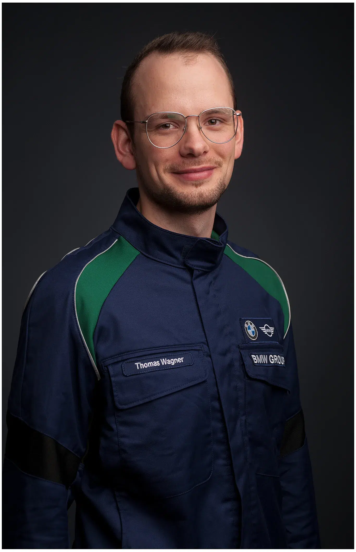 Porträt eines jungen BMW-Ingenieurs in blauer Firmenjacke mit grünen Akzenten und BMW- sowie MINI-Logos, professioneller Gesichtsausdruck.