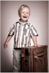 Nachgedacht für einige wenige Sekunden kindermodel-sedcard-belichtungswert-fuer-casting-und-werbung-lachender-junge-mit-altem-koffer-im-fotostudio Kindermodel Sedcard vom Fotostudio Belichtungswert München, lachender blonder Junge im gestreiften Hemd steht neben einem alten braunen Koffer im Studio, geeignet für Kinder-Casting, Werbung und Familienfotografie