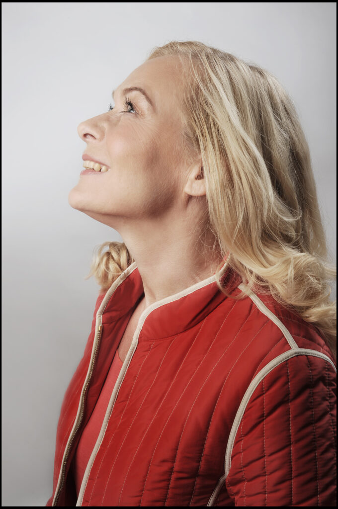Komparsen-Sedcard vom Fotostudio Belichtungswert München, Best Age Blondine lachend im roten Cardigan und gemustertem Kleid, lässig am alten Koffer posierend