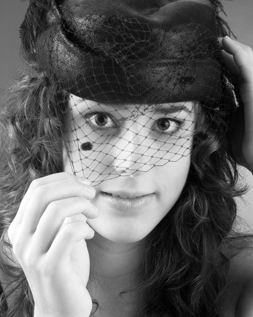 Komparsen Sedcard vom Fotostudio Belichtungswert München, junges Talent im Schwarzweiß-Close-up mit schwarzem Hut, Schleier und Händen am Gesicht