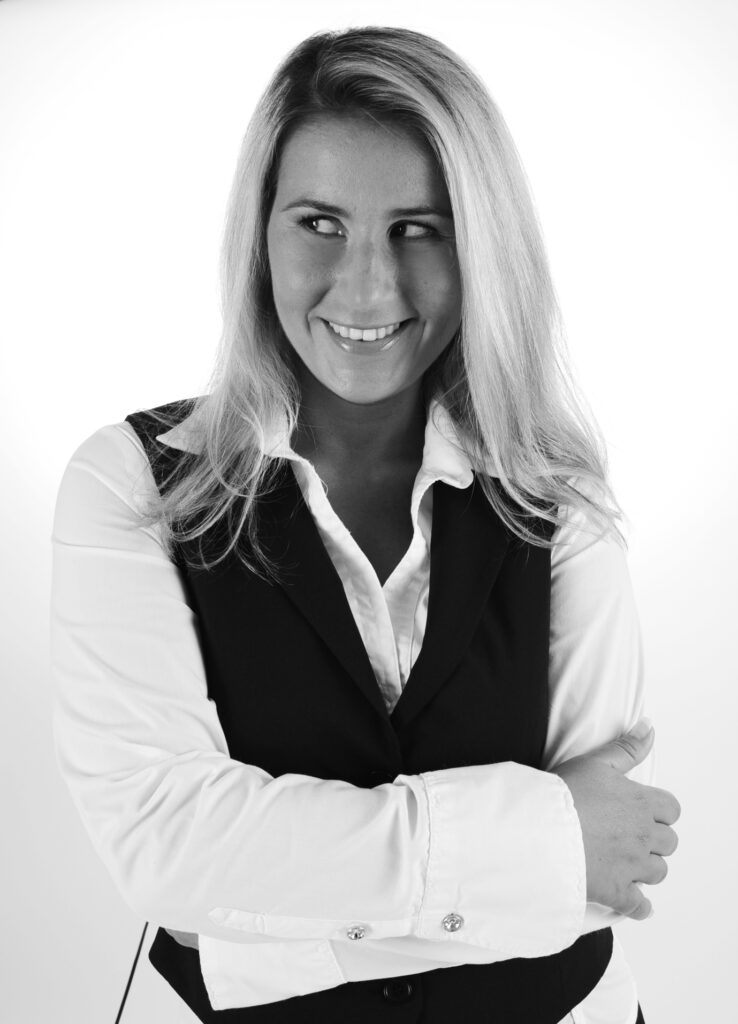 Komparsen-Sedcard vom Fotostudio Belichtungswert München, Tanja im Schwarzweiß-Portrait mit Business-Look, verschränkten Armen, schönen blonden Haaren und seitlichem Lächeln