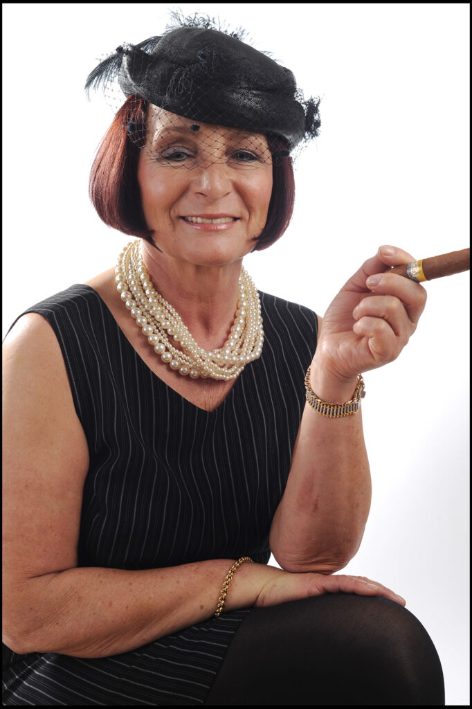 Komparsen Sedcard im Fotostudio Belichtungswert München, Best-Age-Frau mit schwarzem Hut, Perlenkette, Zigarre und witziger Charakterpose für Kleindarstellerrollen