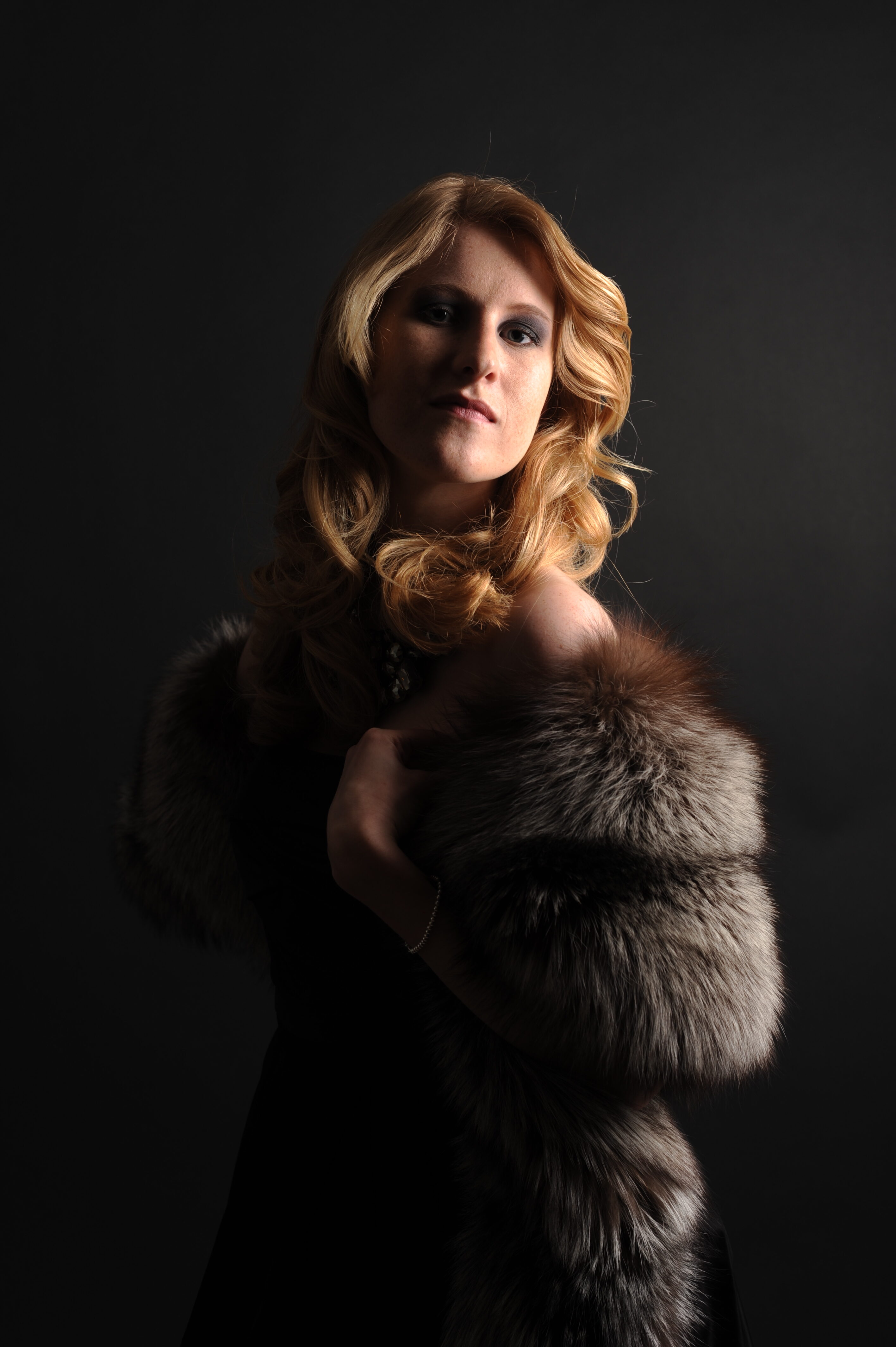 Komparsen Sedcard Fotoshooting im Fotostudio Belichtungswert München, dramatisches Studio-Portrait einer jungen Frau mit blondem Haar, schwarzem Kleid und Fellstola im intensiven Licht