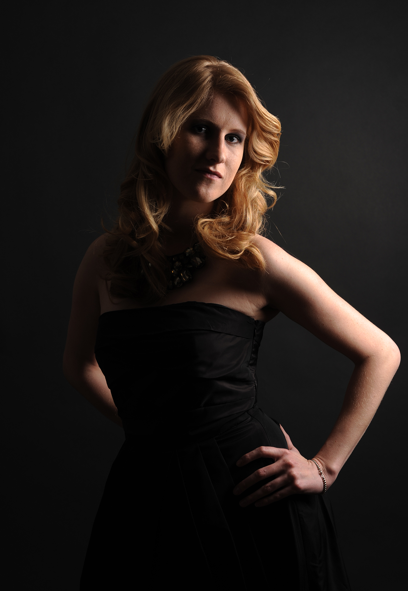 Komparsen Sedcard Fotoshooting im Fotostudio Belichtungswert München, junge blonde Frau in schwarzem Kleid mit intensivem Blick und Hand in der Taille vor dunklem Studiohintergrund