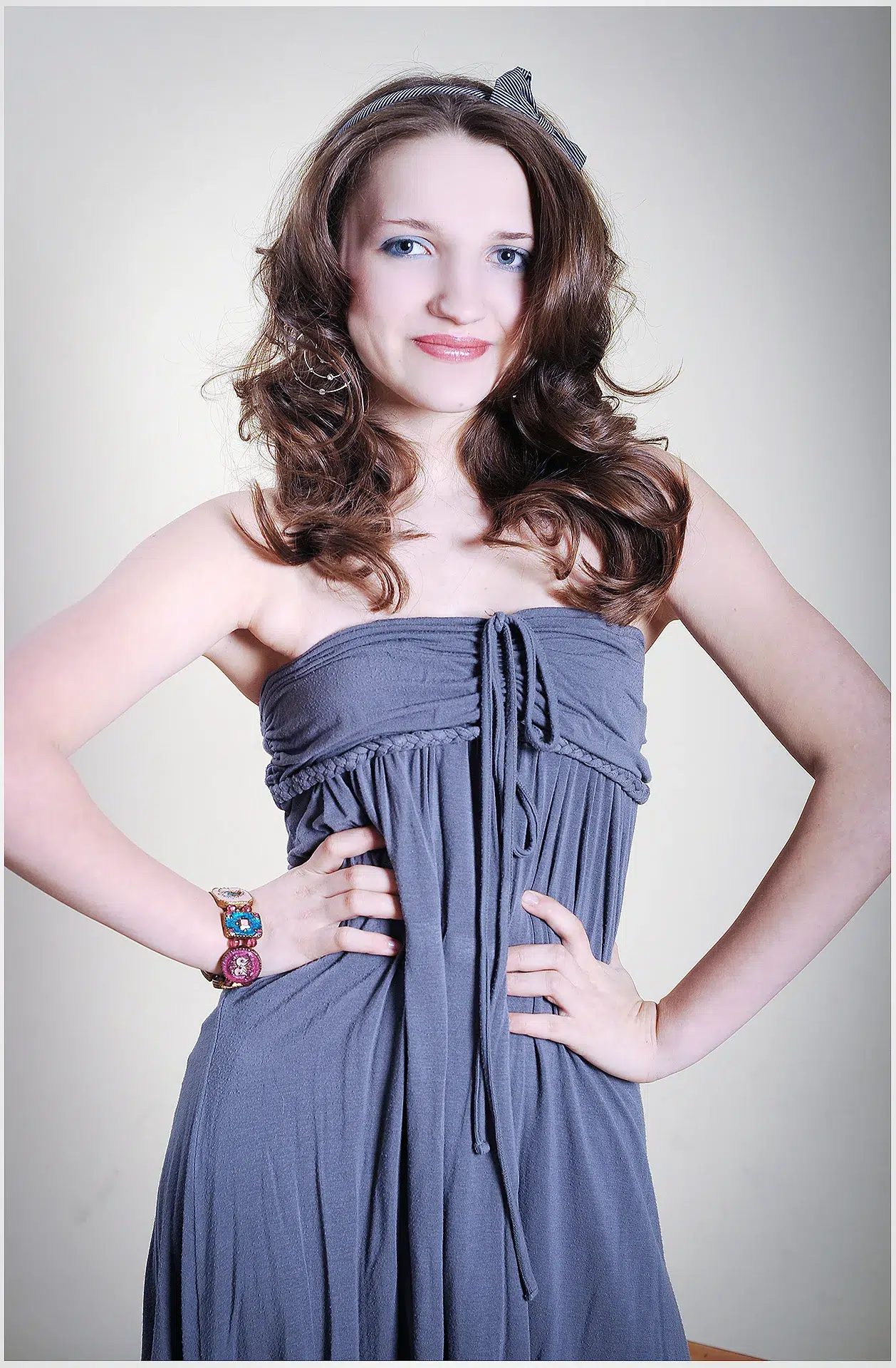 Ein junges Mädchen mit langen, voluminösen Locken trägt ein elegantes graues Kleid mit einem gerafften Oberteil und schulterfreien Ärmeln. Ihre Hände ruhen selbstbewusst in der Taille, während sie mit einem strahlenden Lächeln in die Kamera blickt. Eine dezente Haarspange hält ihr Haar zurück, und eine bunte Armbanduhr verleiht ihrem Look eine verspielte Note. Der Hintergrund ist hell und neutral, wodurch das Model im Mittelpunkt steht.

