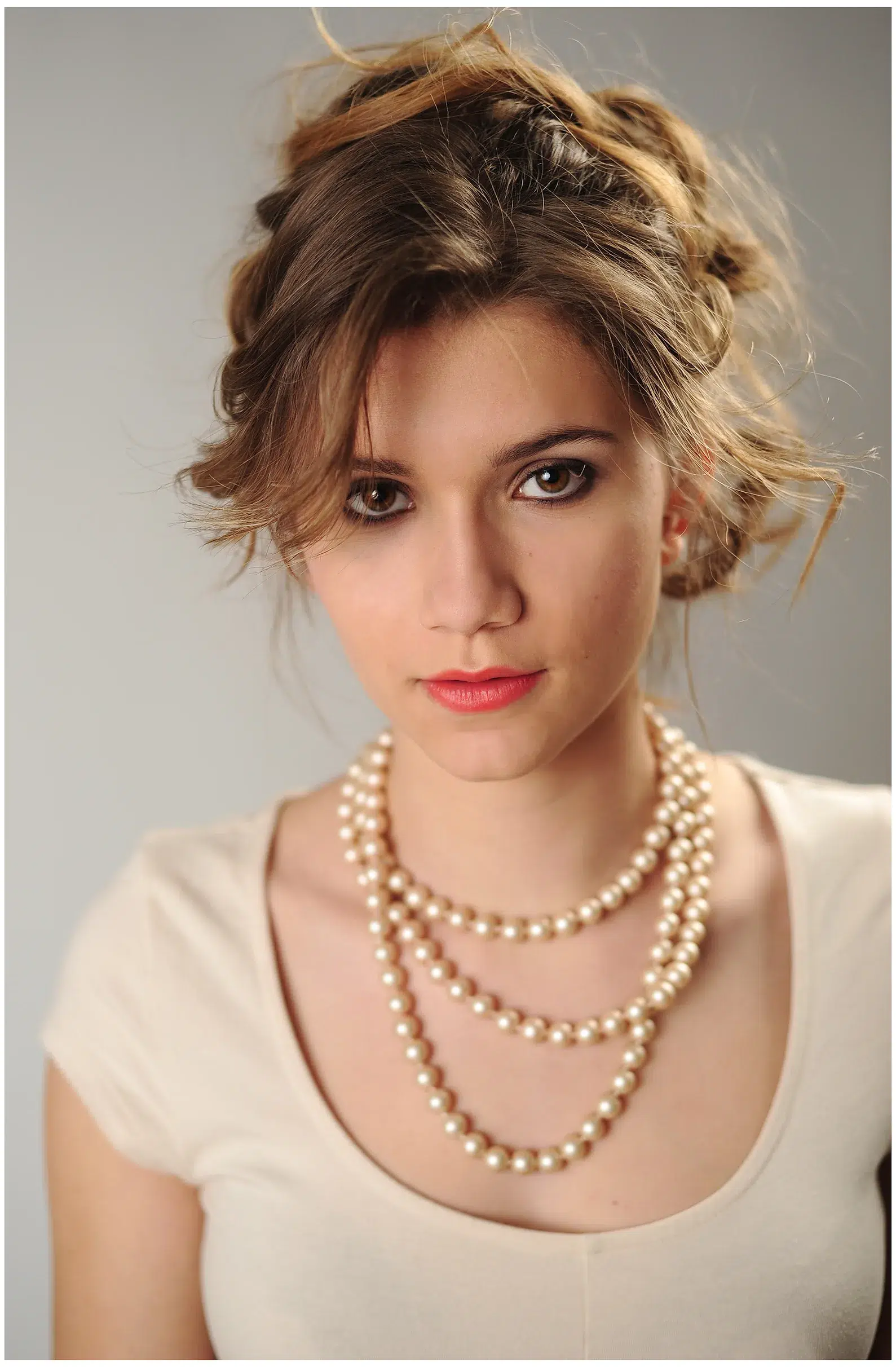 Ein junges Mädchen mit eleganter Hochsteckfrisur trägt ein schlichtes, cremefarbenes Oberteil und eine mehrreihige Perlenkette. Ihr Blick ist ruhig und intensiv, ihre Lippen sind sanft geschminkt. Die Beleuchtung ist weich und schmeichelhaft, wodurch ihr Gesicht sanft modelliert wird. Der Hintergrund ist dezent grau, was den Fokus auf ihre feinen Gesichtszüge lenkt.








