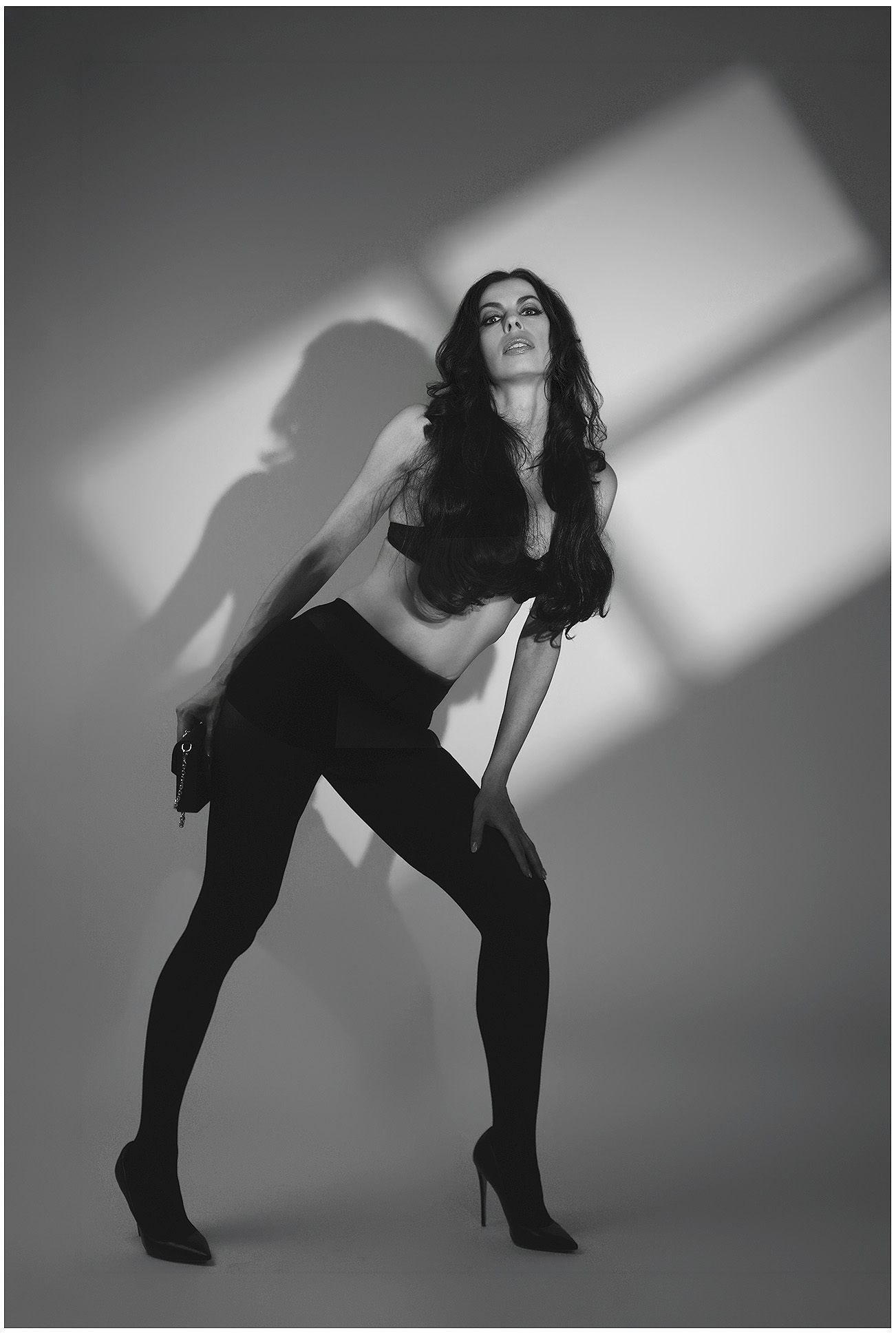 Schwarzweiß-Ganzkörperportrait aus einem Model-Fotoshooting in München: Frau mit langen dunklen Haaren, schwarzem Fashion-Outfit und High Heels posiert vor dramatischen Licht- und Schattenflächen.