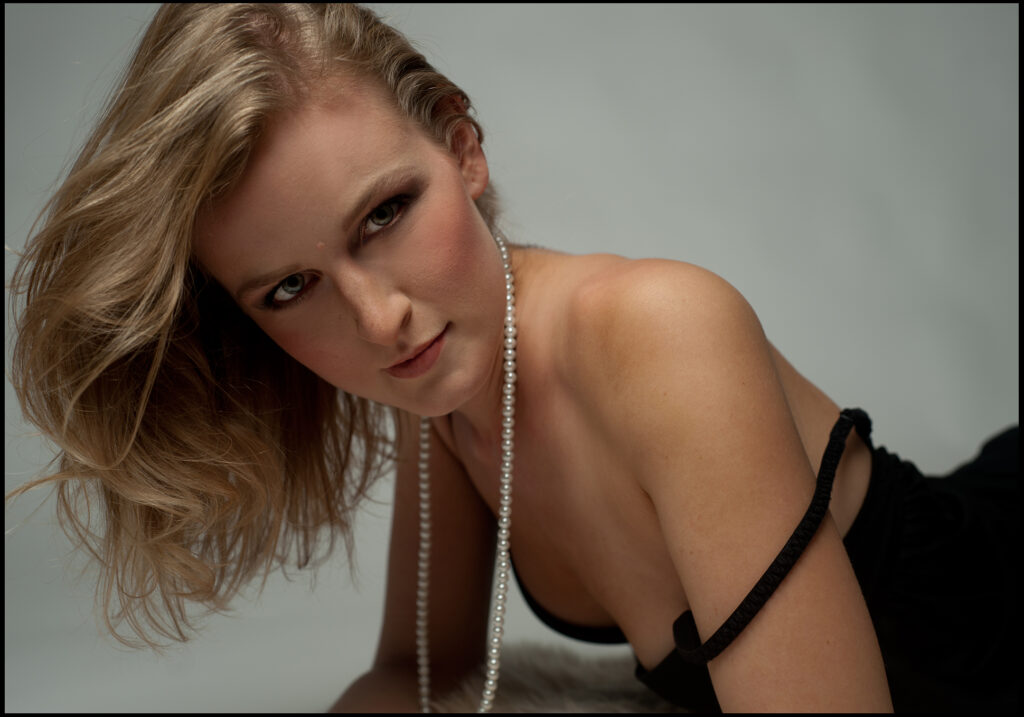 Junges Anfängermodel beim Sedcard-Fotoshooting im Fotostudio Belichtungswert München, liegende Fashion-Pose mit Perlenkette, blondem Haar und intensivem direktem Blick