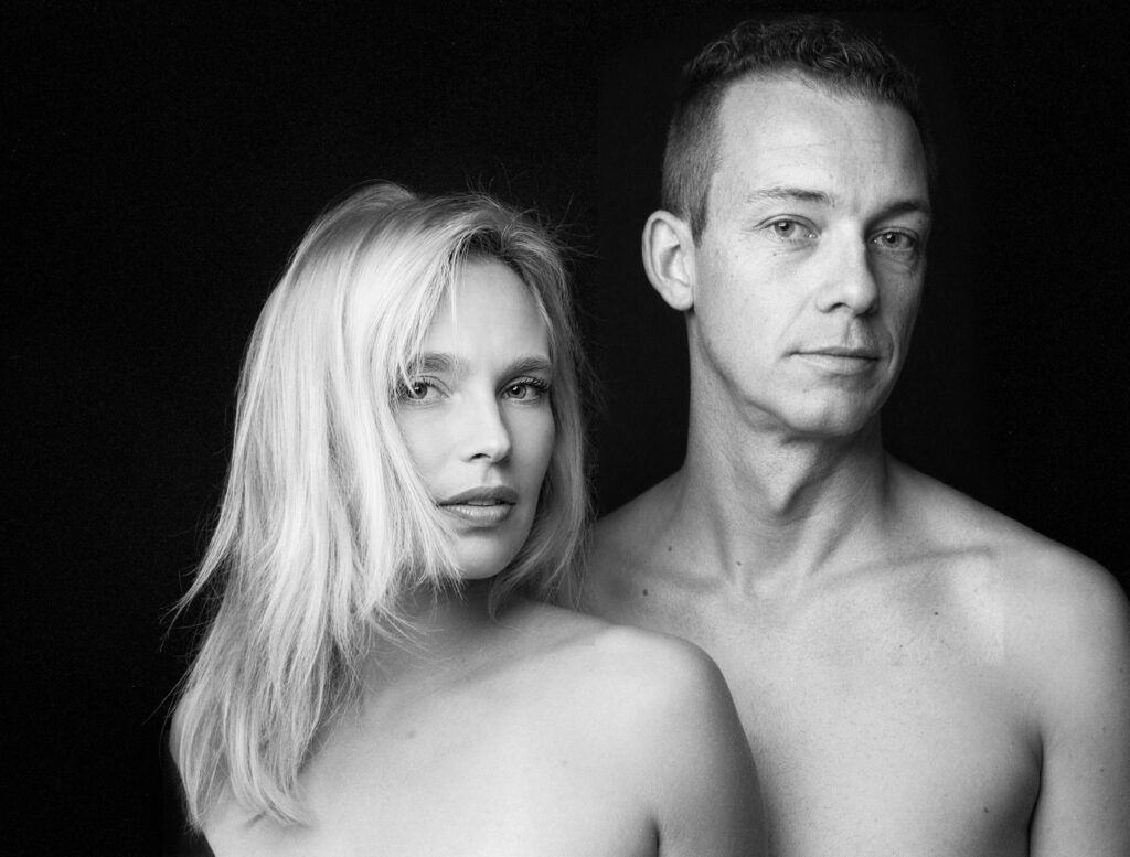 Schwarz-Weiß-Portrait eines Paares beim Paarshooting, Couple-Fotoshooting im Fotostudio München, blonde Frau und Mann mit freien Schultern vor schwarzem Hintergrund, nah und ausdrucksstark fotografiert